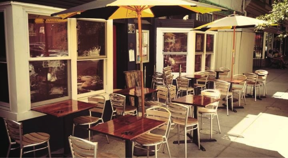 Outdoor sidewalk seating with wooden tables, metal chairs, and yellow and white umbrellas lining a sunny urban block in soft daylight