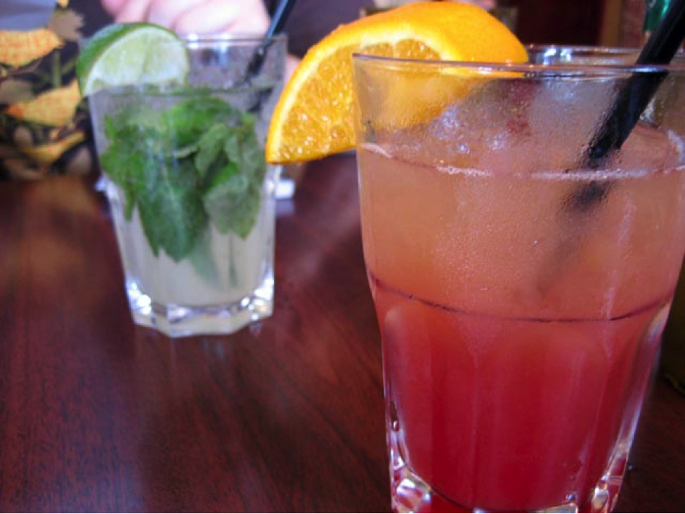 Two beverages in glasses on a wooden table, one reddish cocktail with orange garnish and straw, another with lime and mint, relaxed atmosphere