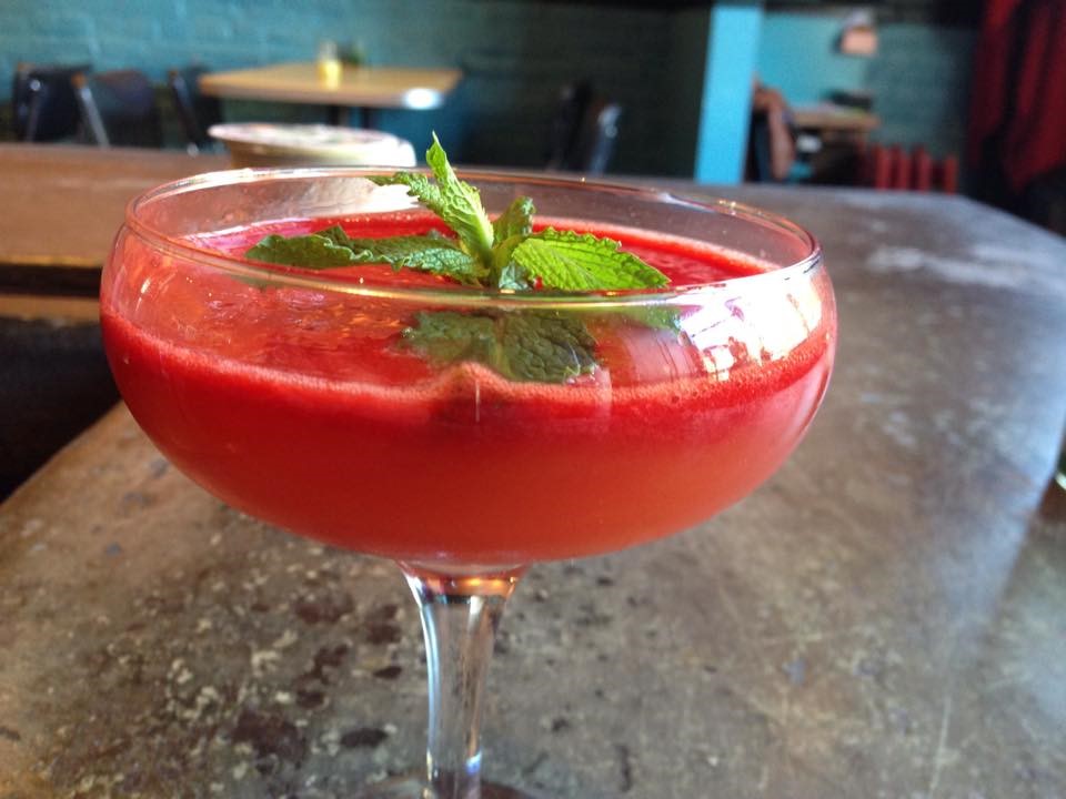 Close-up of a vivid red cocktail in a coupe glass garnished with mint leaves, resting on a warm wooden bar in deep crimson and green tones