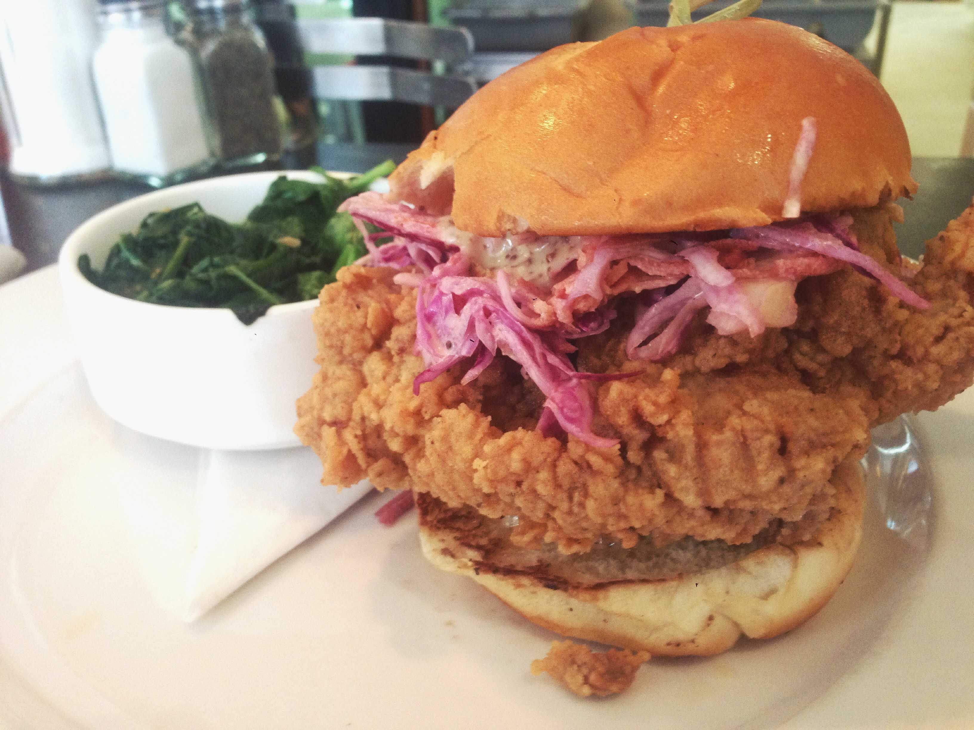 Close-up of a fried chicken sandwich with purple slaw on a glossy bun beside a bowl of sautéed greens, in warm rust and green tones