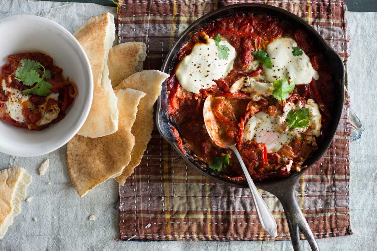 Overhead shot of a cast-iron skillet with a tomato and egg dish garnished with cilantro, served alongside pita bread on a wooden table